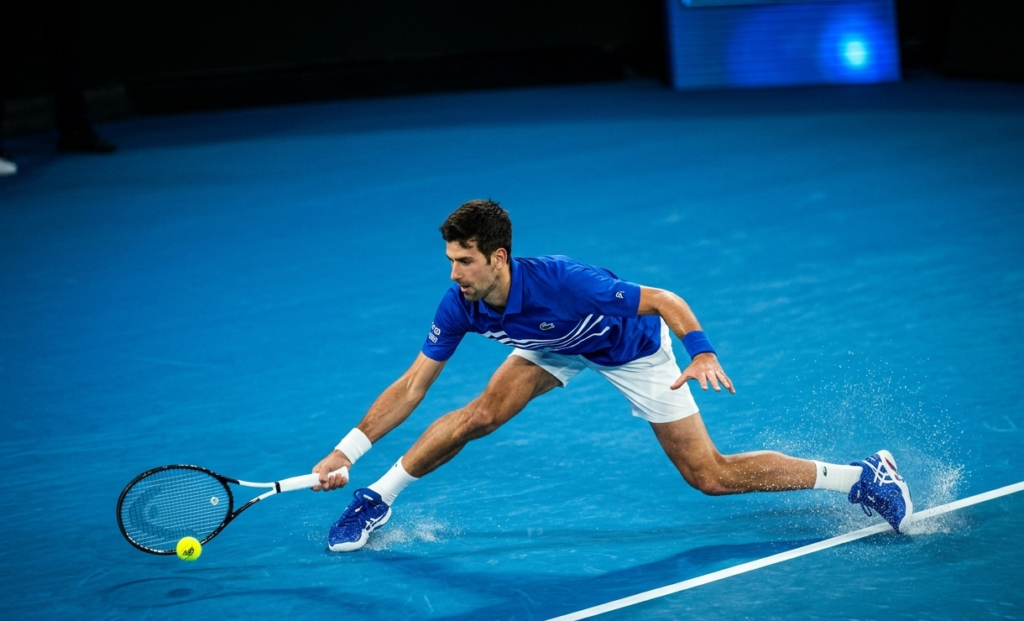 Alt Text: A dynamic sports photograph of a male tennis player (Novak Djokovic) stretching across a vibrant blue hard court to hit a groundstroke.